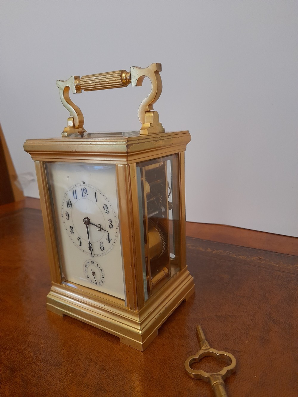 A gilt brass carriage clock featuring a fluted top handle, bevelled glass panels, a white enamel main dial with Arabic numerals, an alarm sub-dial, and an accompanying winding key. View III