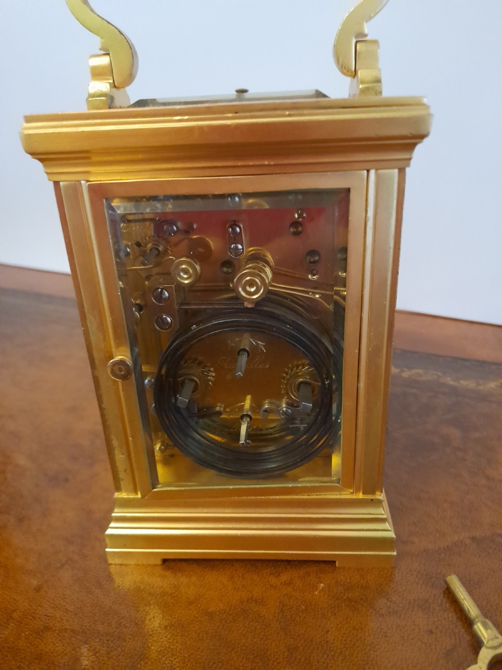 A gilt brass carriage clock featuring a fluted top handle, bevelled glass panels, a white enamel main dial with Arabic numerals, an alarm sub-dial, and an accompanying winding key. View VII