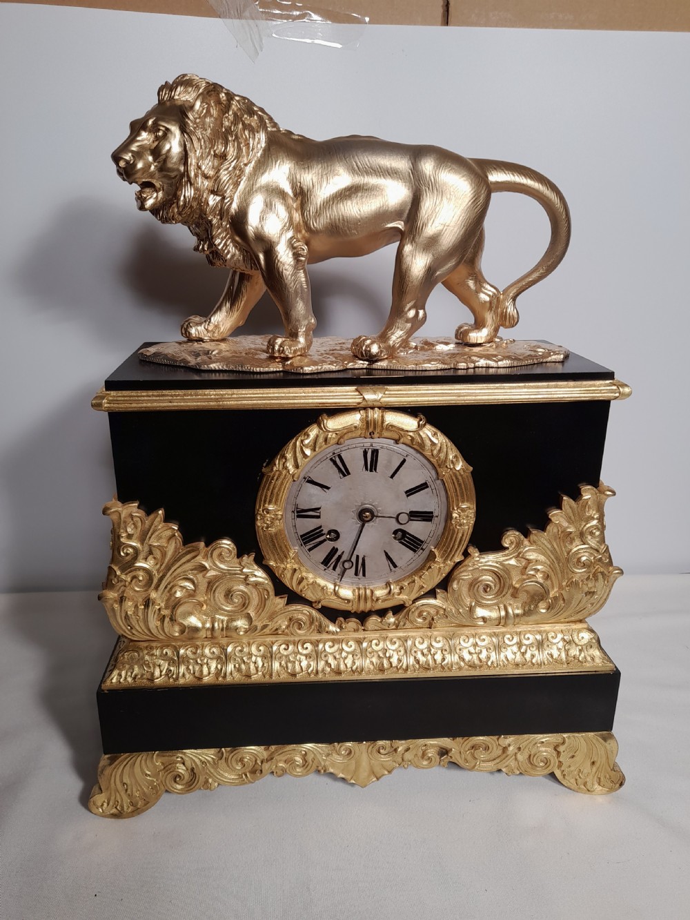 A French Empire style mantel clock featuring a gilded lion sculpture on top of a black rectangular casing adorned with gold acanthus mounts and a central clock dial.