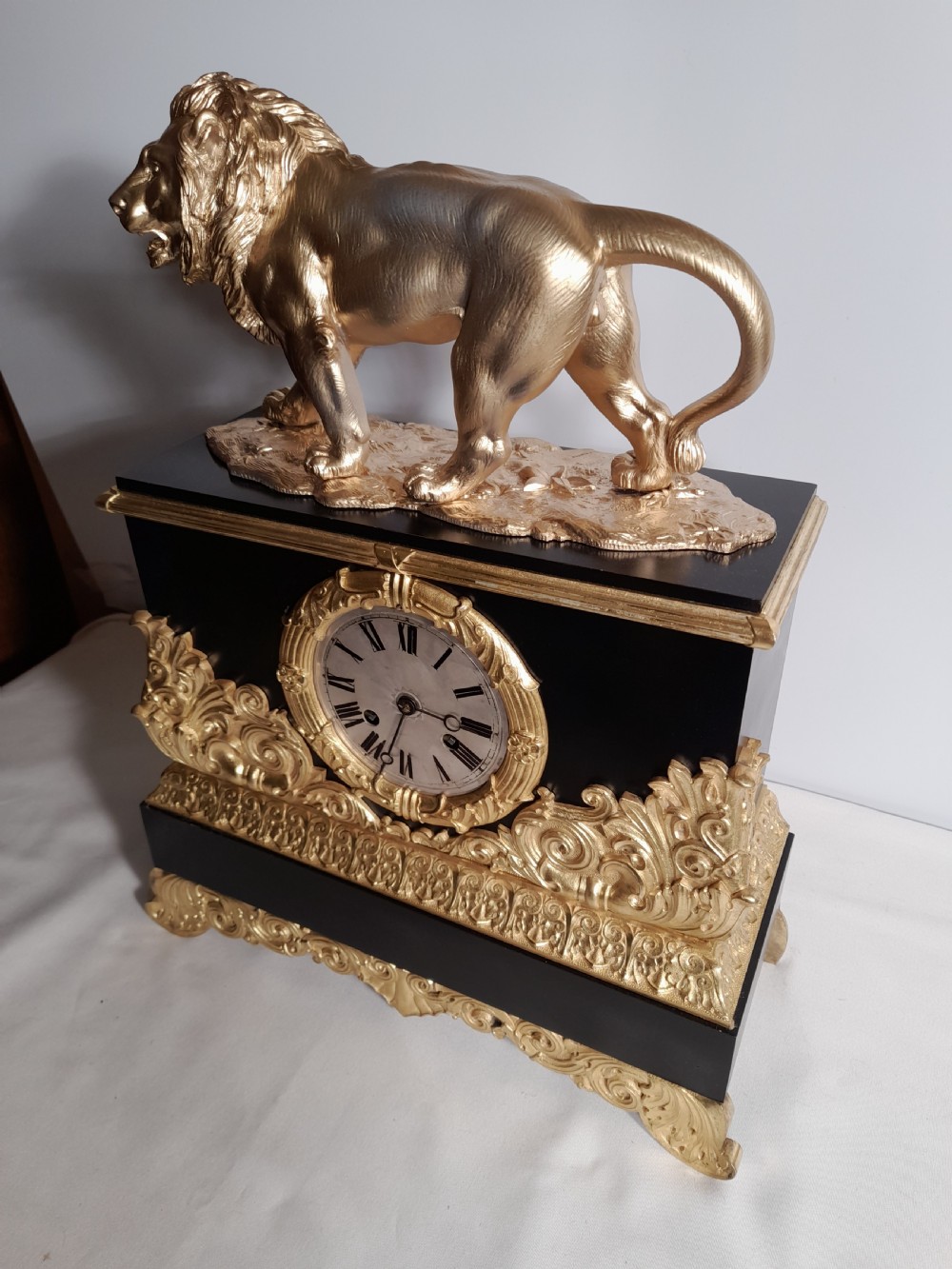 A French Empire style mantel clock featuring a gilded lion sculpture on top of a black rectangular casing adorned with gold acanthus mounts and a central clock dial. View II