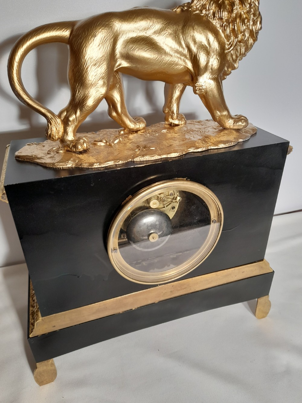 A French Empire style mantel clock featuring a gilded lion sculpture on top of a black rectangular casing adorned with gold acanthus mounts and a central clock dial. View V