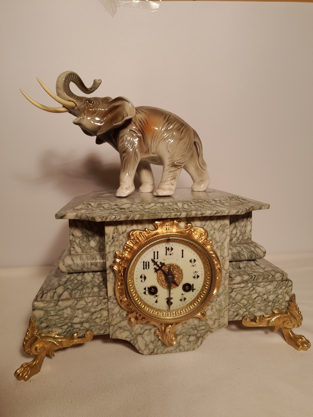 An antique mantel clock crafted from veined green marble with decorative gilt mounts and a glazed ceramic elephant figure resting on top.