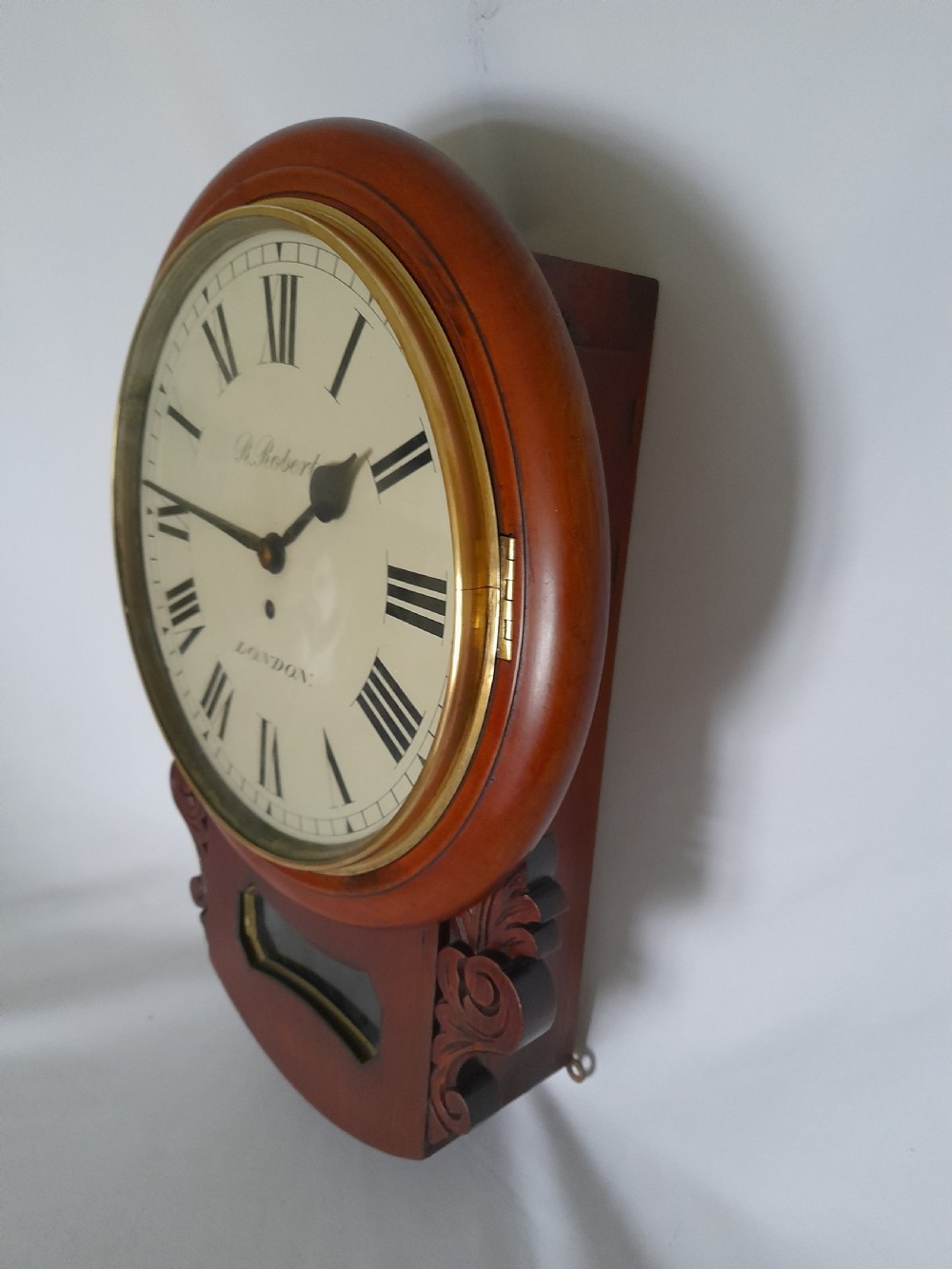 A Victorian mahogany drop-dial wall clock featuring a brass-rimmed cream dial with Roman numerals, black spade hands, and carved scrollwork details on the lower case.