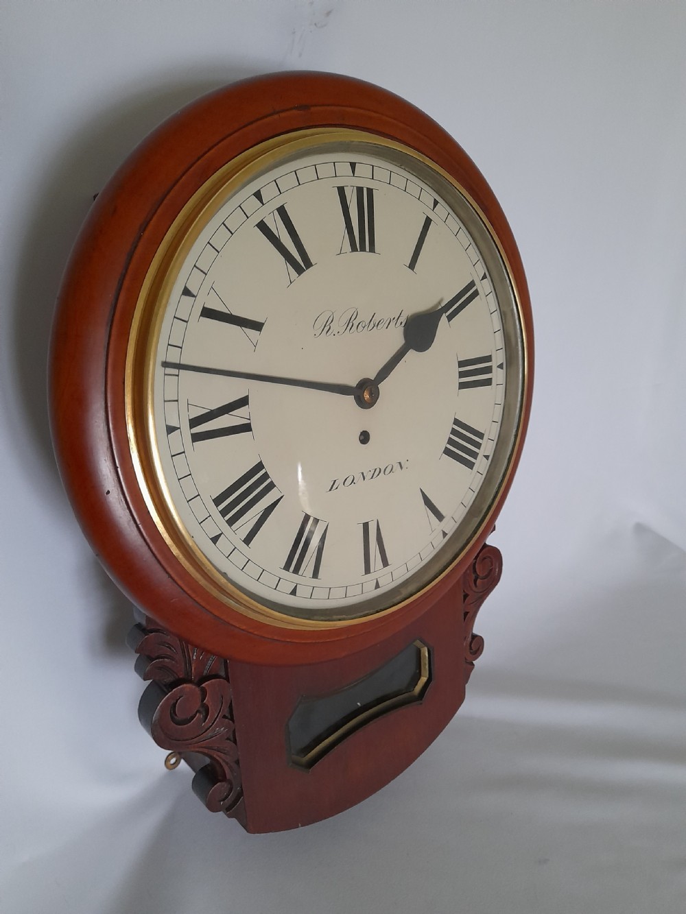 A Victorian mahogany drop-dial wall clock featuring a brass-rimmed cream dial with Roman numerals, black spade hands, and carved scrollwork details on the lower case. View II