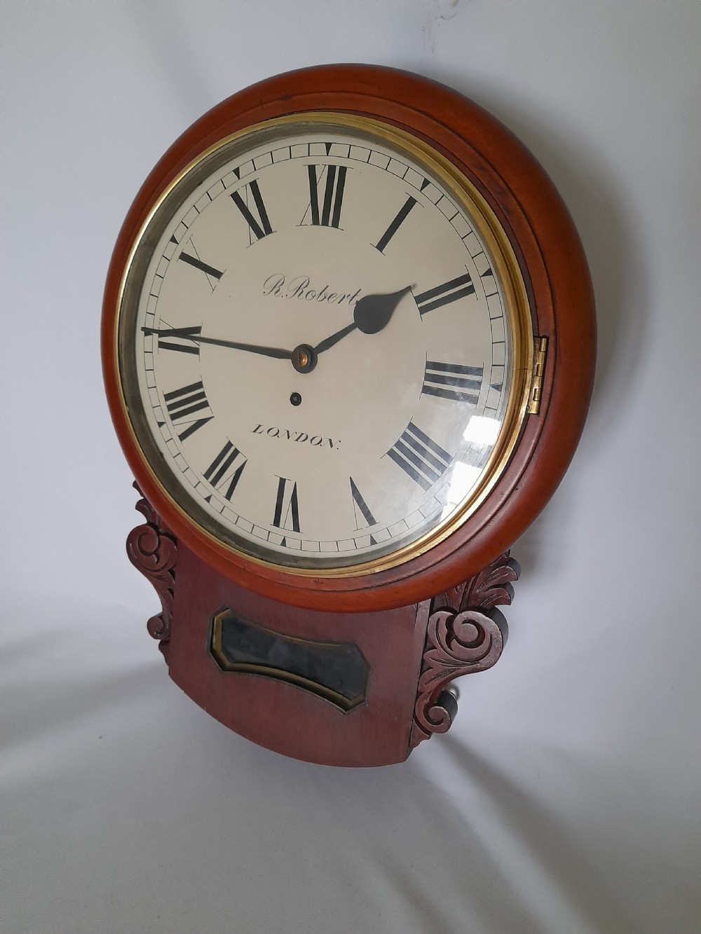 A Victorian mahogany drop-dial wall clock featuring a brass-rimmed cream dial with Roman numerals, black spade hands, and carved scrollwork details on the lower case. View III