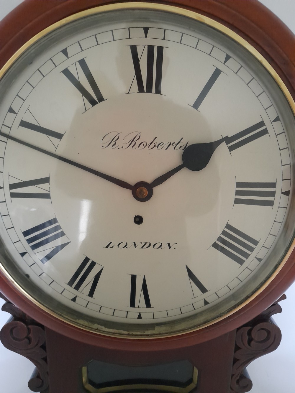 A Victorian mahogany drop-dial wall clock featuring a brass-rimmed cream dial with Roman numerals, black spade hands, and carved scrollwork details on the lower case. View IV