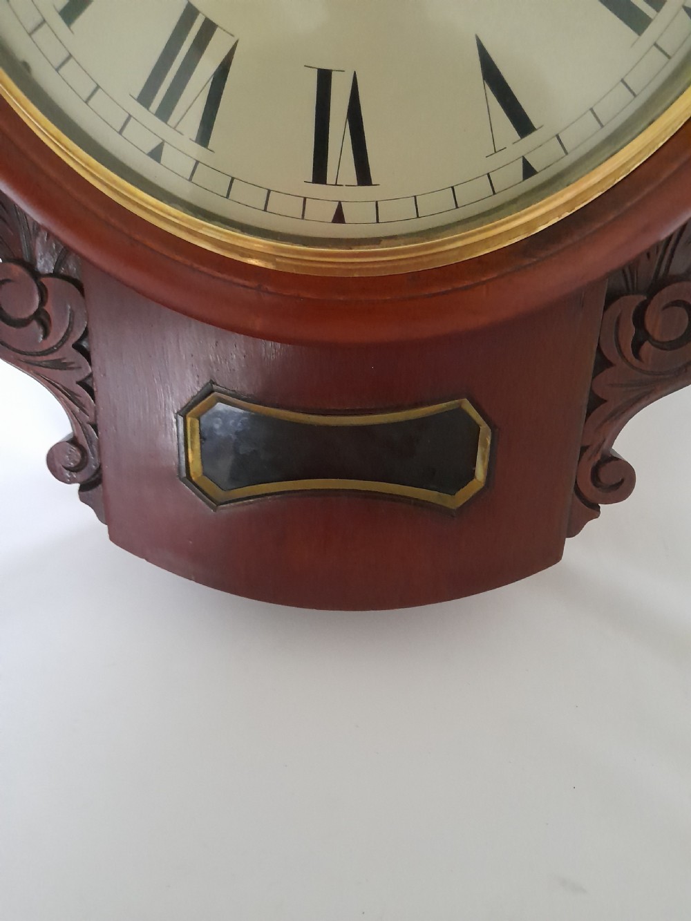 A Victorian mahogany drop-dial wall clock featuring a brass-rimmed cream dial with Roman numerals, black spade hands, and carved scrollwork details on the lower case. View V