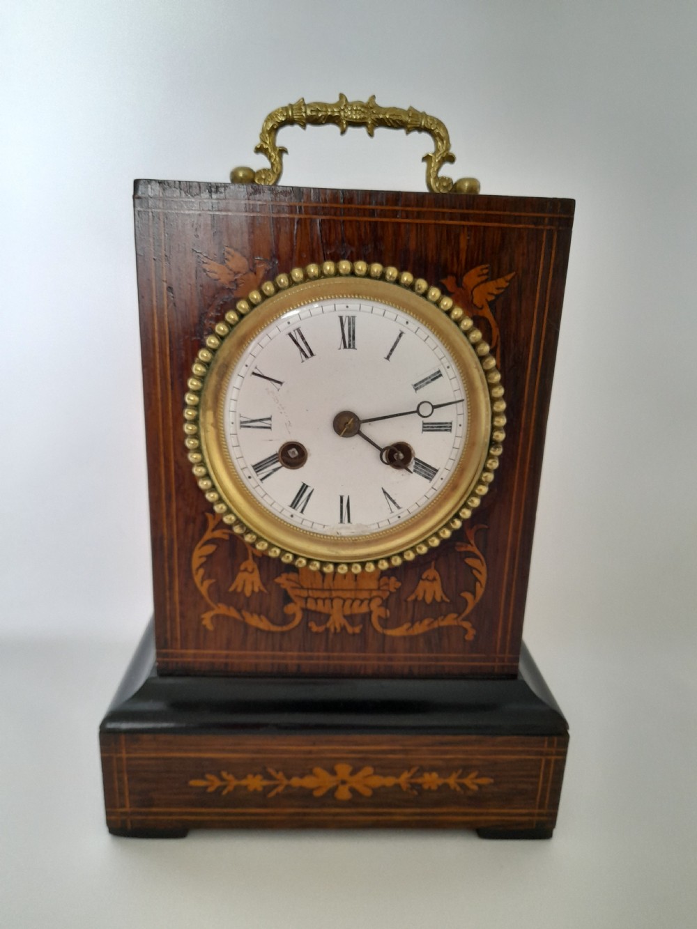 An antique French officers clock with a dark wood inlaid rectangular case, a decorative brass carrying handle, and a white enamel clock face with Roman numerals.