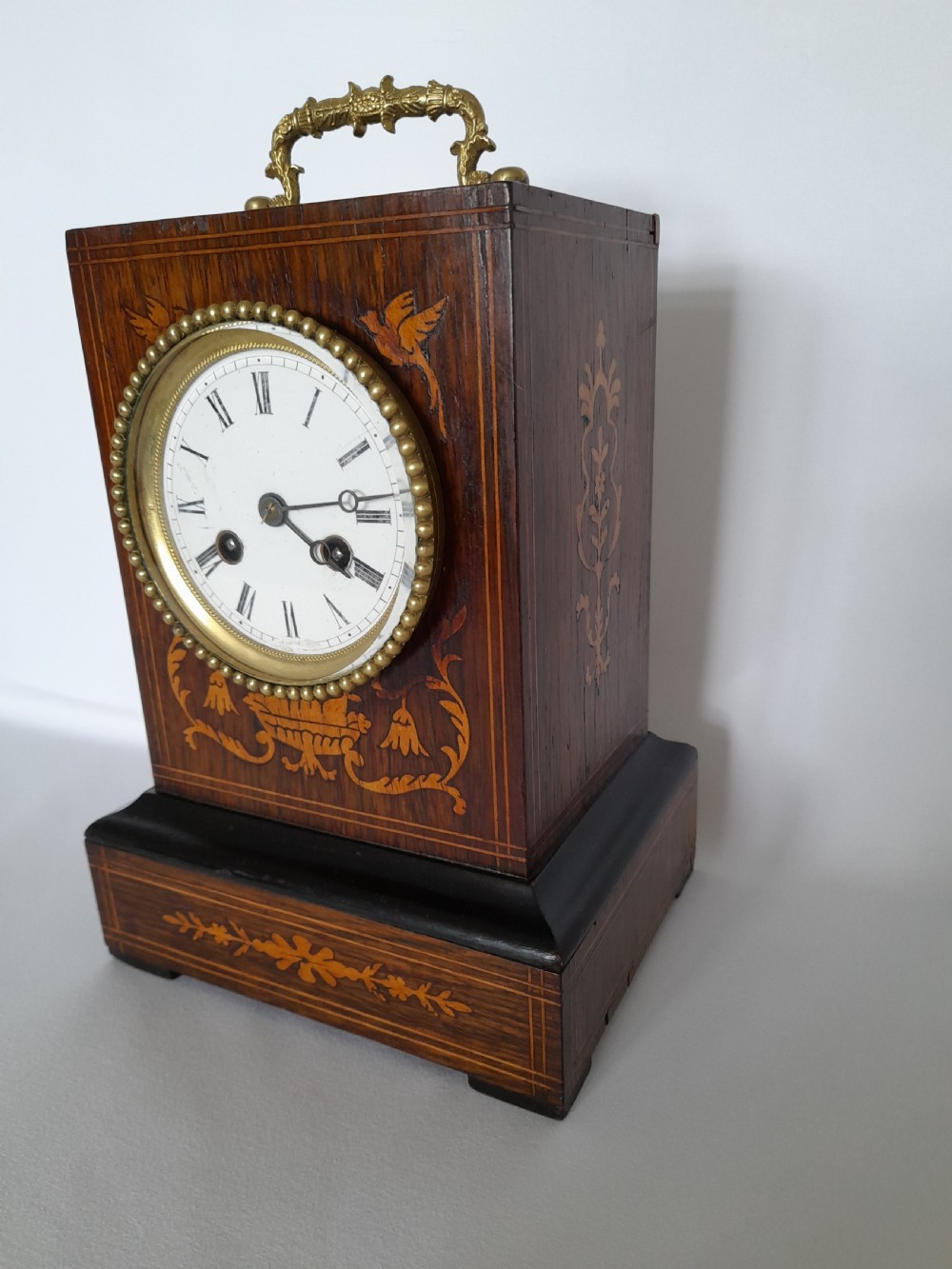 An antique French officers clock with a dark wood inlaid rectangular case, a decorative brass carrying handle, and a white enamel clock face with Roman numerals. View II