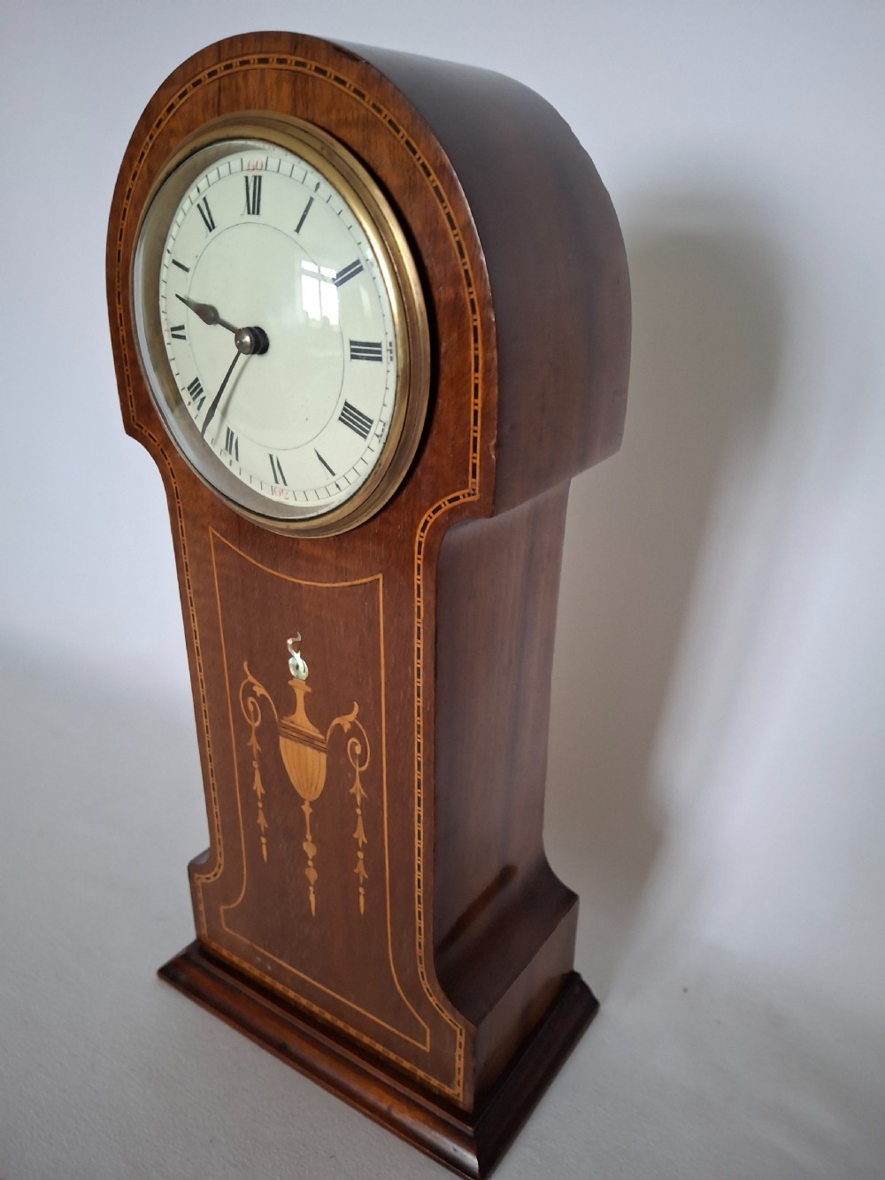 An Art Nouveau mahogany mantel clock featuring a balloon-shaped case with inlaid urn marquetry, boxwood stringing, a circular white dial with Roman numerals, and a brass bezel. View X