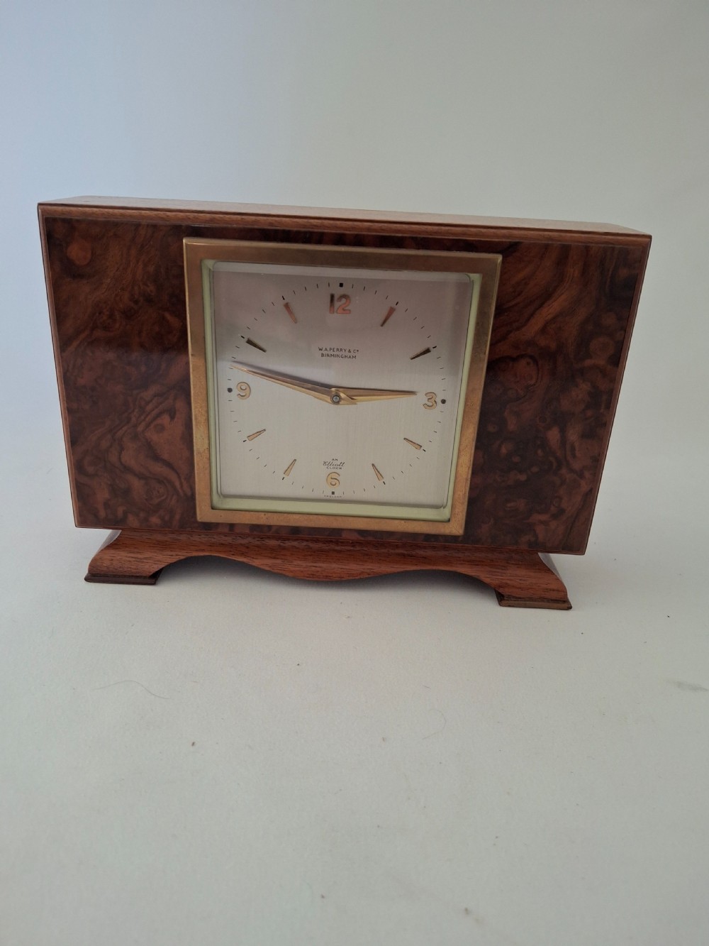 A rectangular burr walnut desk clock featuring a square brass bezel, silvered dial, and gold hands on a shaped plinth base.