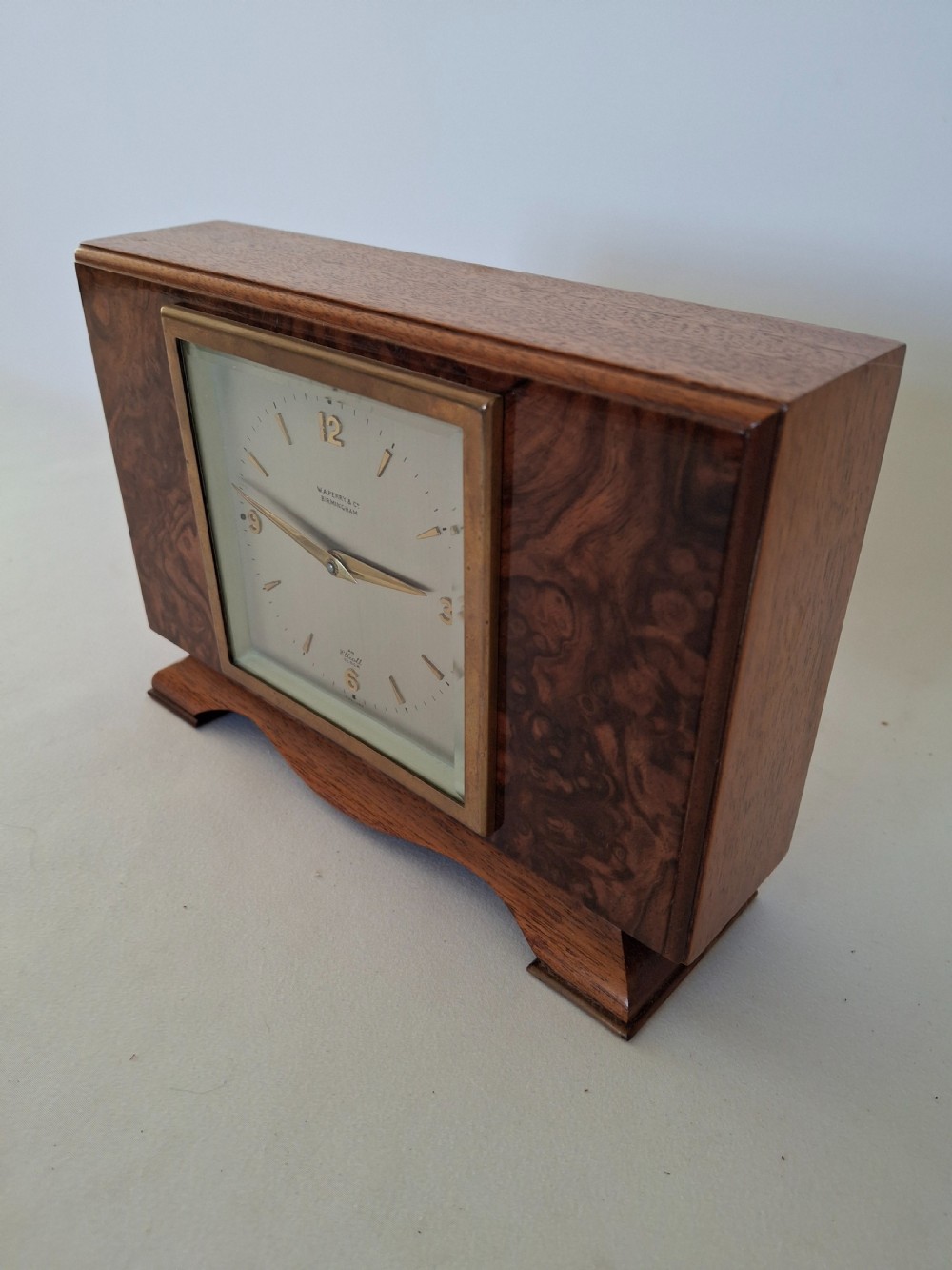 A rectangular burr walnut desk clock featuring a square brass bezel, silvered dial, and gold hands on a shaped plinth base. View II