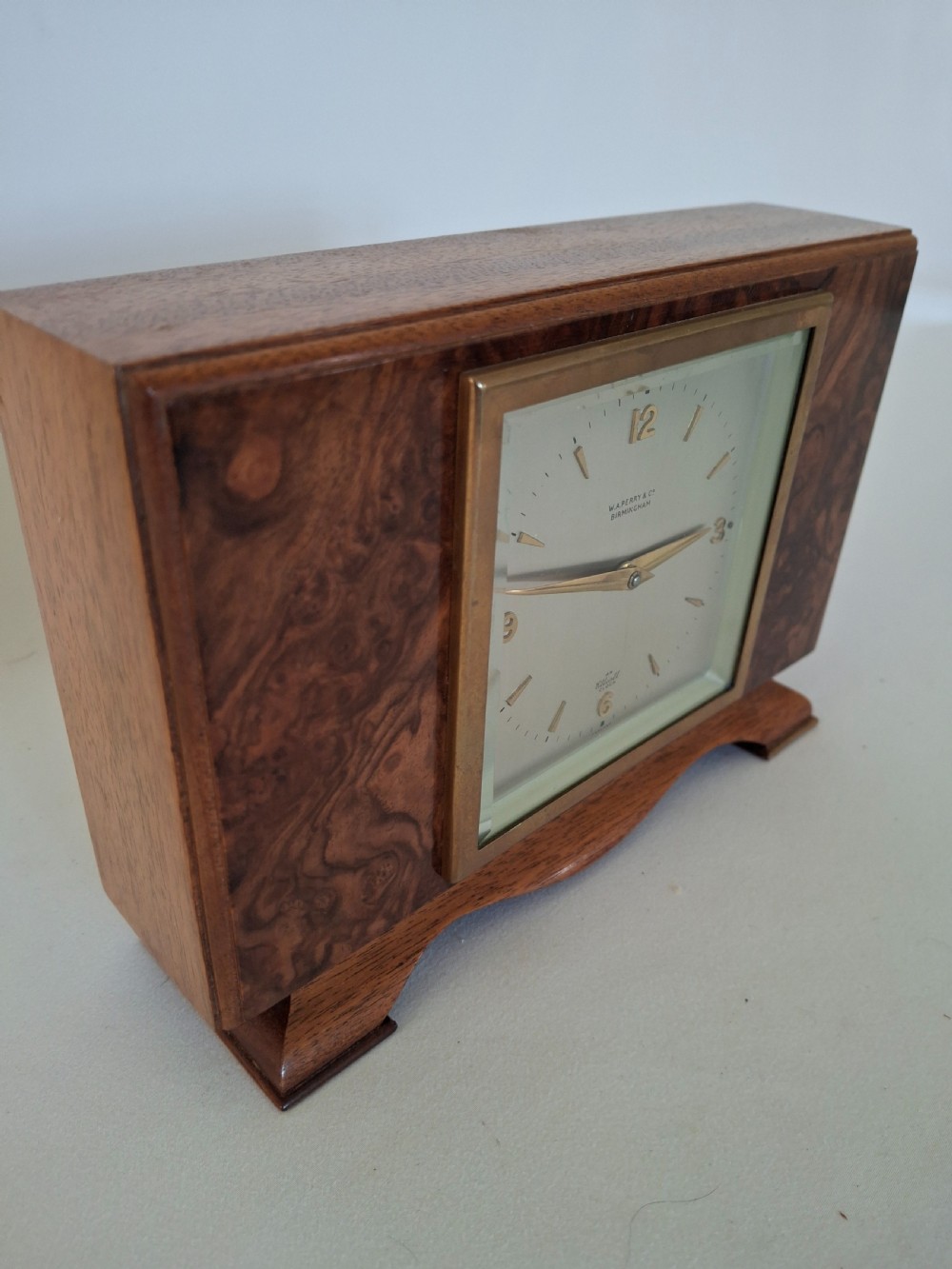 A rectangular burr walnut desk clock featuring a square brass bezel, silvered dial, and gold hands on a shaped plinth base. View III