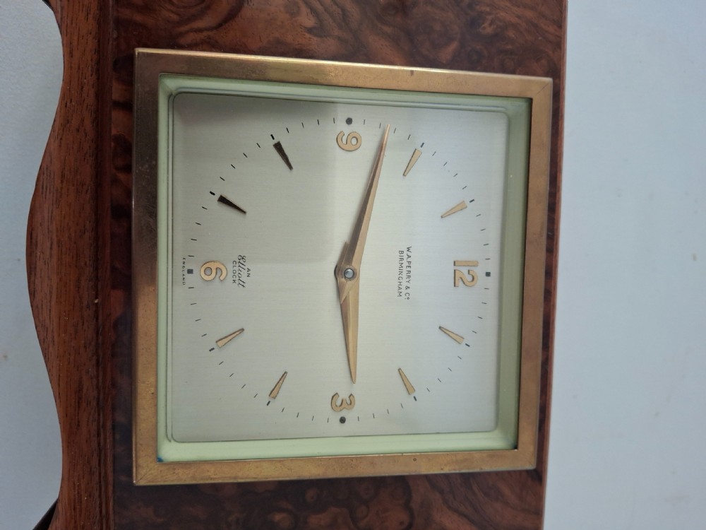 A rectangular burr walnut desk clock featuring a square brass bezel, silvered dial, and gold hands on a shaped plinth base. View V
