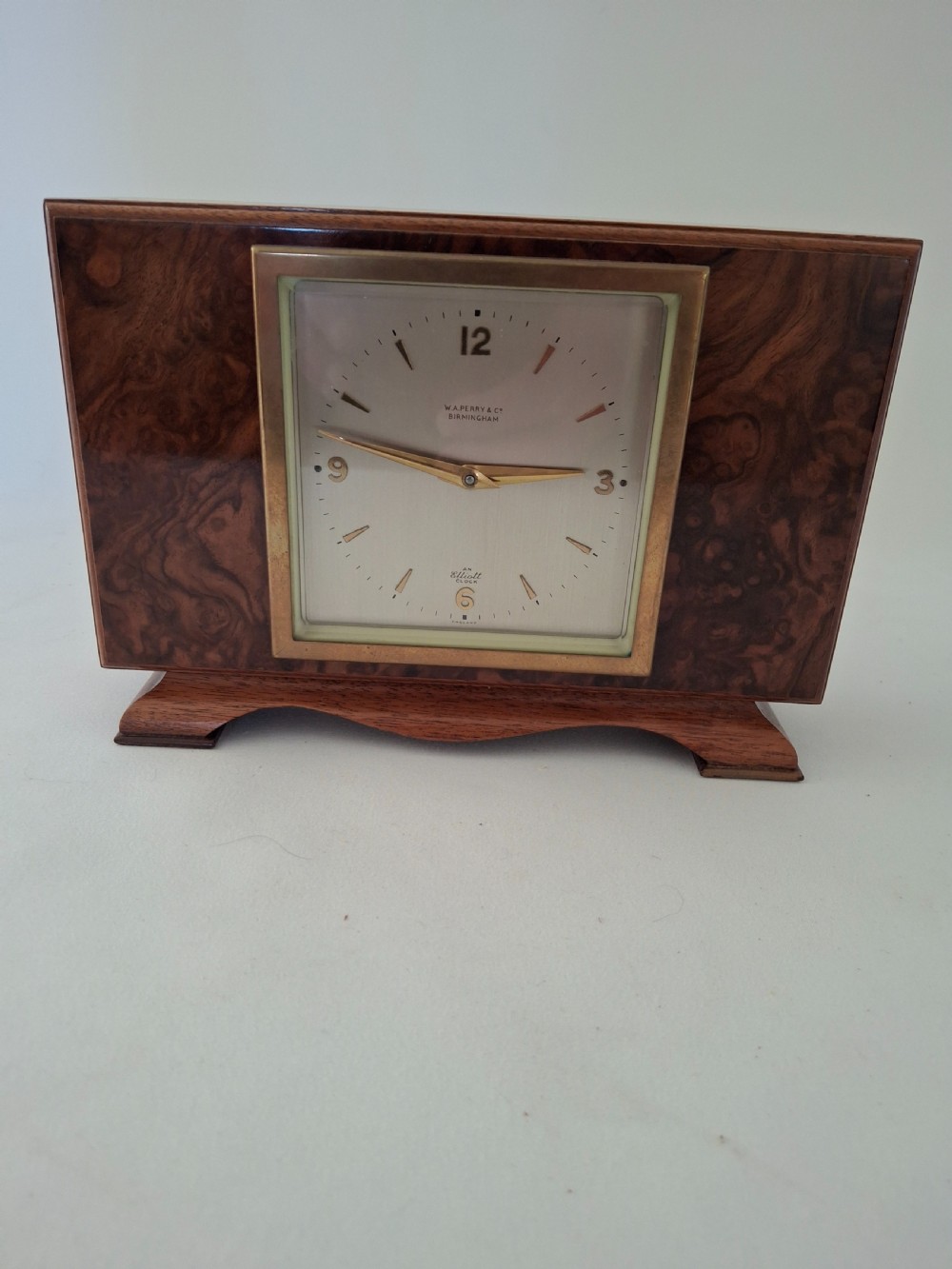 A rectangular burr walnut desk clock featuring a square brass bezel, silvered dial, and gold hands on a shaped plinth base. View VI