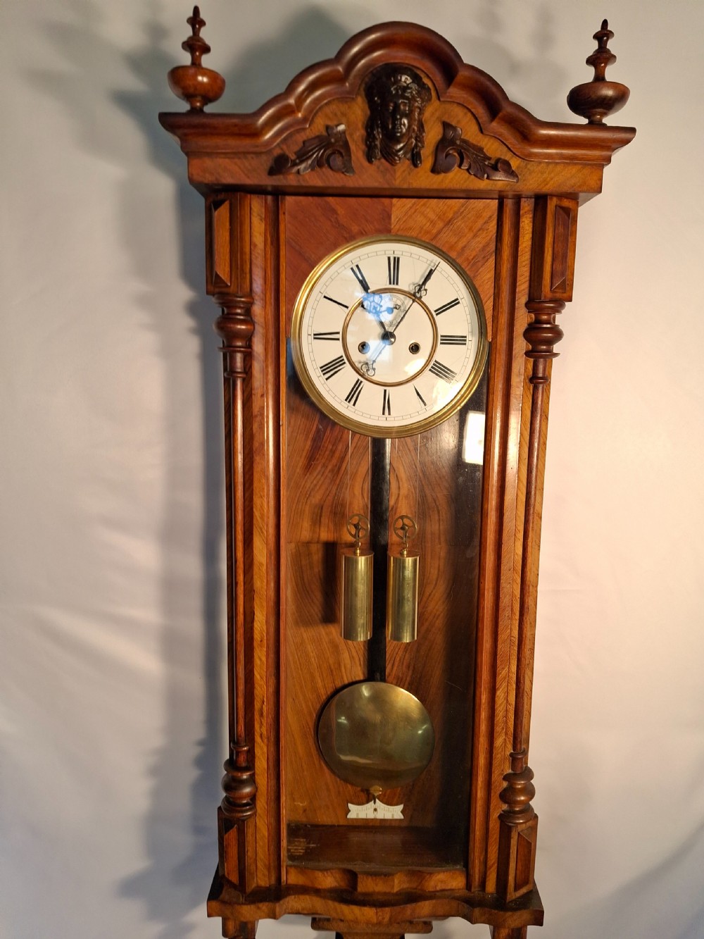 An antique walnut double-weight Vienna regulator wall clock featuring a white enamel dial with Roman numerals, visible brass weights, and a brass pendulum inside a glazed case with turned finials.