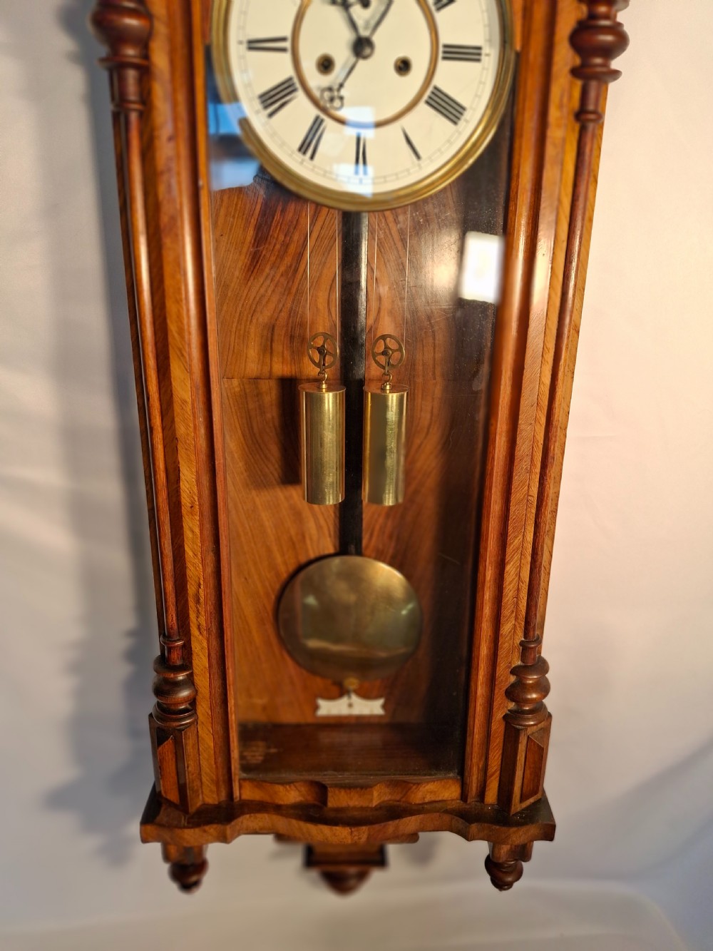 An antique walnut double-weight Vienna regulator wall clock featuring a white enamel dial with Roman numerals, visible brass weights, and a brass pendulum inside a glazed case with turned finials. View III