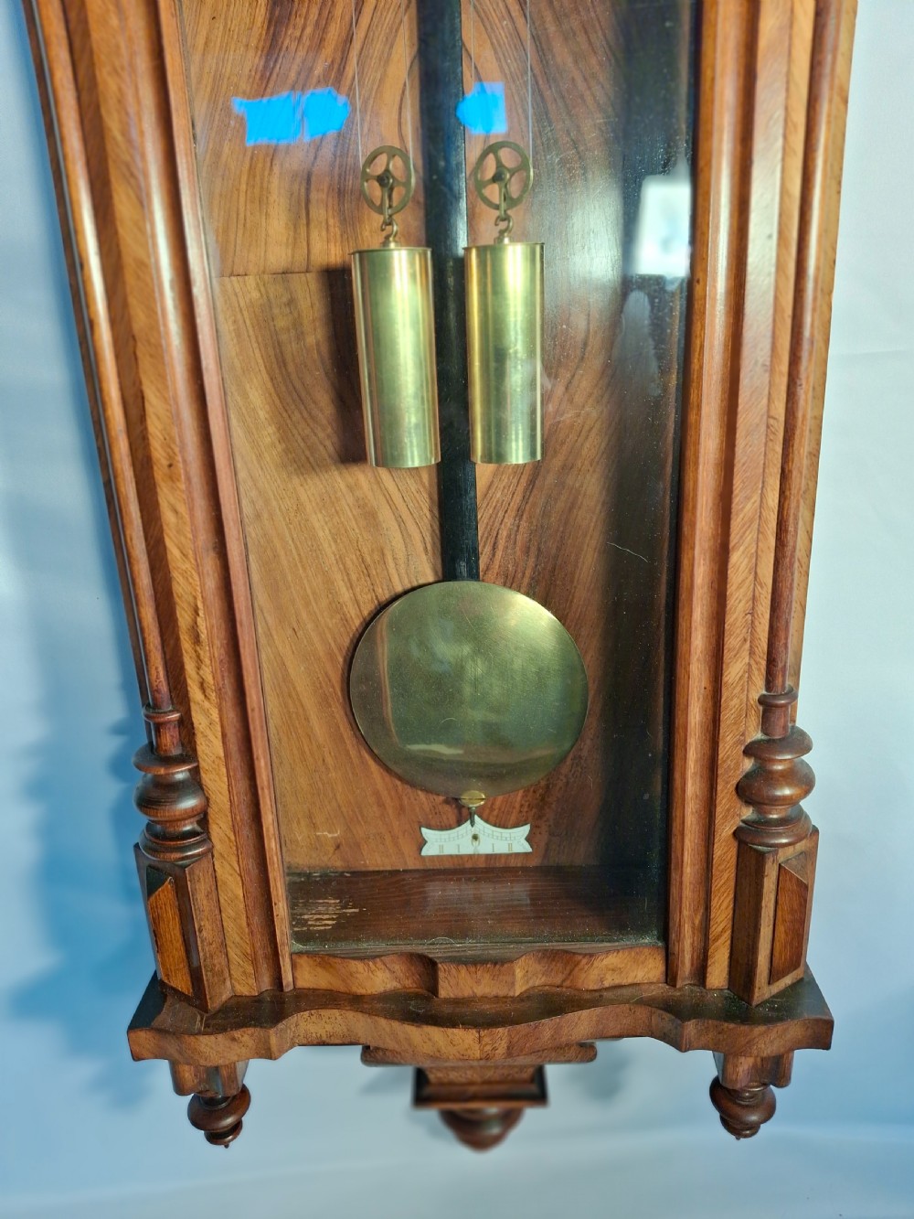An antique walnut double-weight Vienna regulator wall clock featuring a white enamel dial with Roman numerals, visible brass weights, and a brass pendulum inside a glazed case with turned finials. View VI