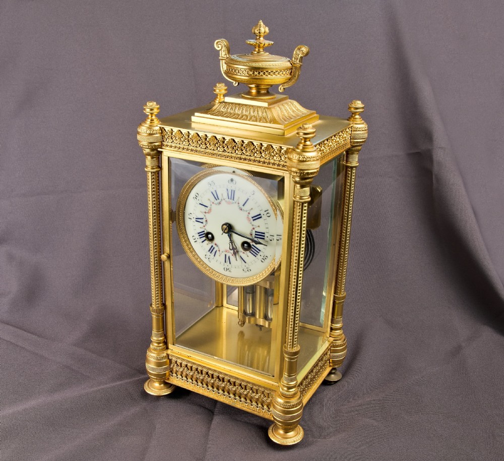 A French Neoclassical gilt brass four-glass mantel clock featuring an urn finial, fluted corner columns, an enamel dial with Roman numerals, and a visible mercury pendulum.