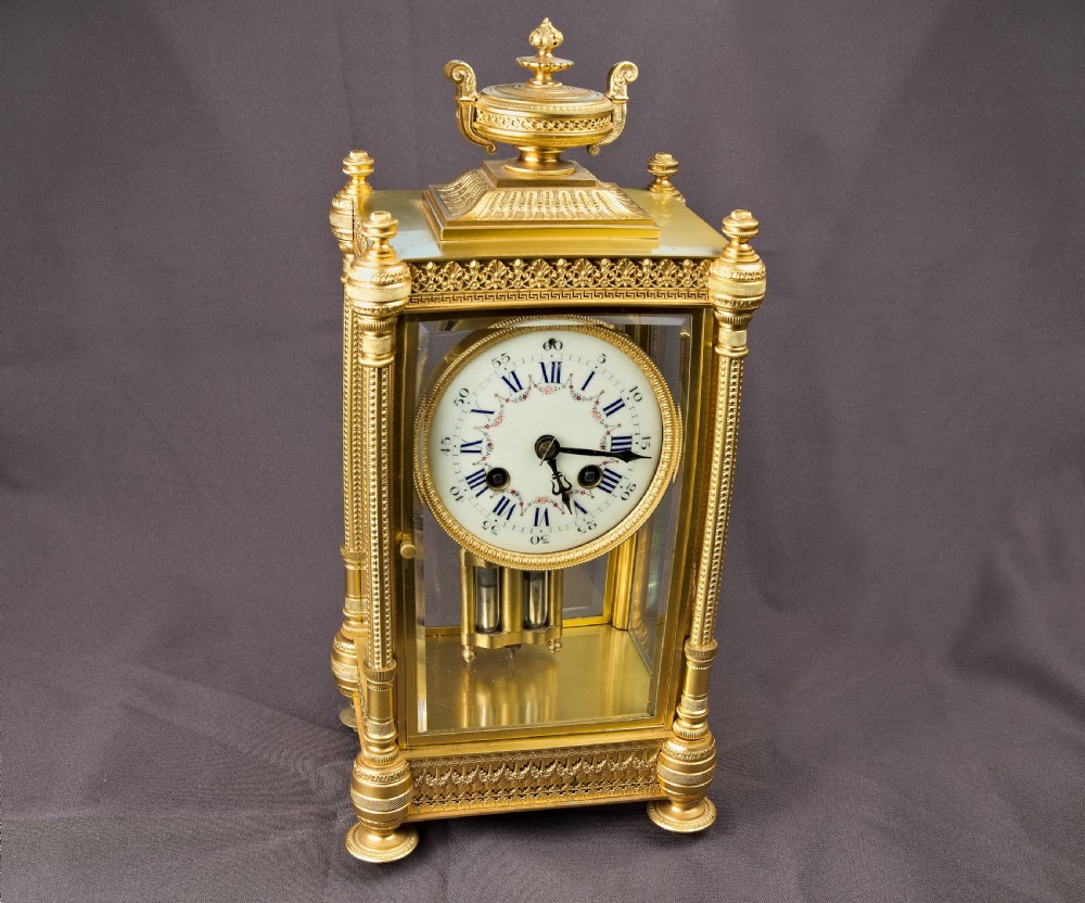 A French Neoclassical gilt brass four-glass mantel clock featuring an urn finial, fluted corner columns, an enamel dial with Roman numerals, and a visible mercury pendulum. View II