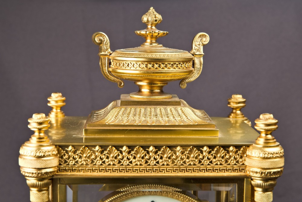 A French Neoclassical gilt brass four-glass mantel clock featuring an urn finial, fluted corner columns, an enamel dial with Roman numerals, and a visible mercury pendulum. View III