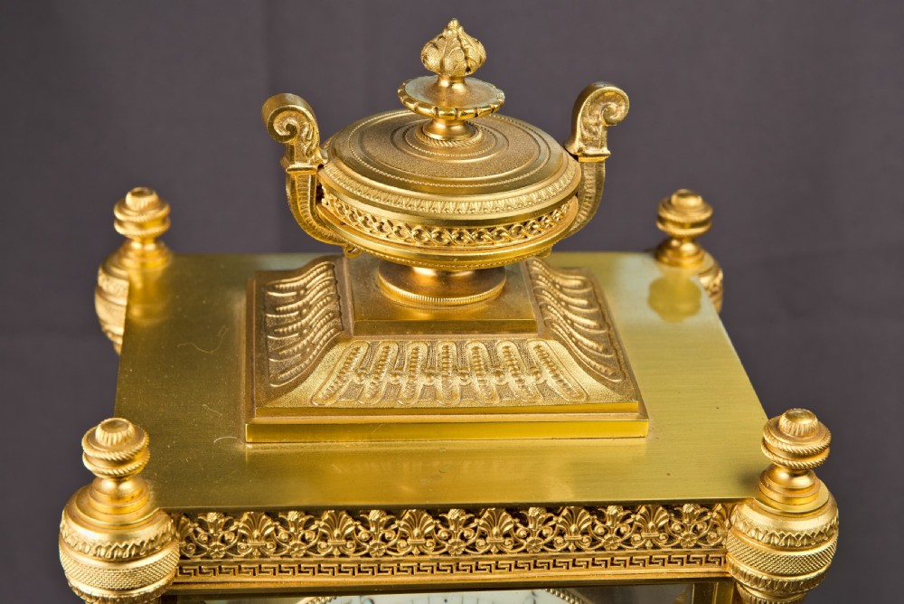 A French Neoclassical gilt brass four-glass mantel clock featuring an urn finial, fluted corner columns, an enamel dial with Roman numerals, and a visible mercury pendulum. View V