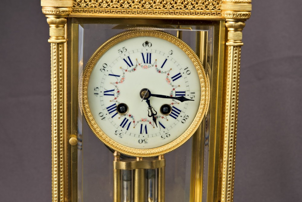 A French Neoclassical gilt brass four-glass mantel clock featuring an urn finial, fluted corner columns, an enamel dial with Roman numerals, and a visible mercury pendulum. View VI