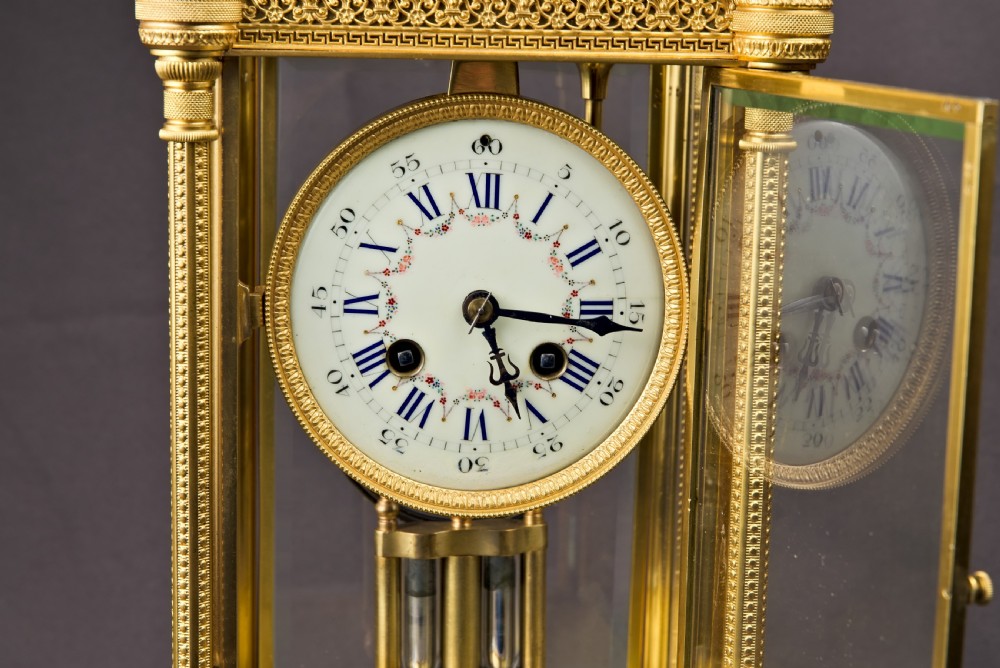A French Neoclassical gilt brass four-glass mantel clock featuring an urn finial, fluted corner columns, an enamel dial with Roman numerals, and a visible mercury pendulum. View VII
