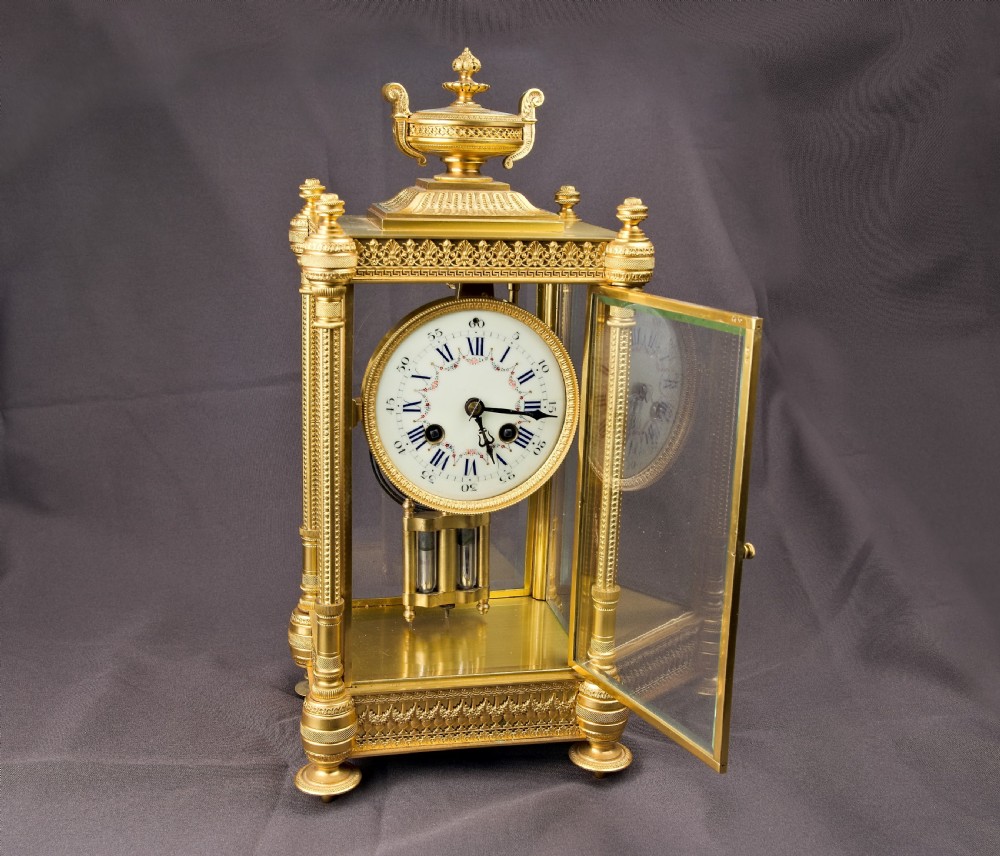 A French Neoclassical gilt brass four-glass mantel clock featuring an urn finial, fluted corner columns, an enamel dial with Roman numerals, and a visible mercury pendulum. View VIII