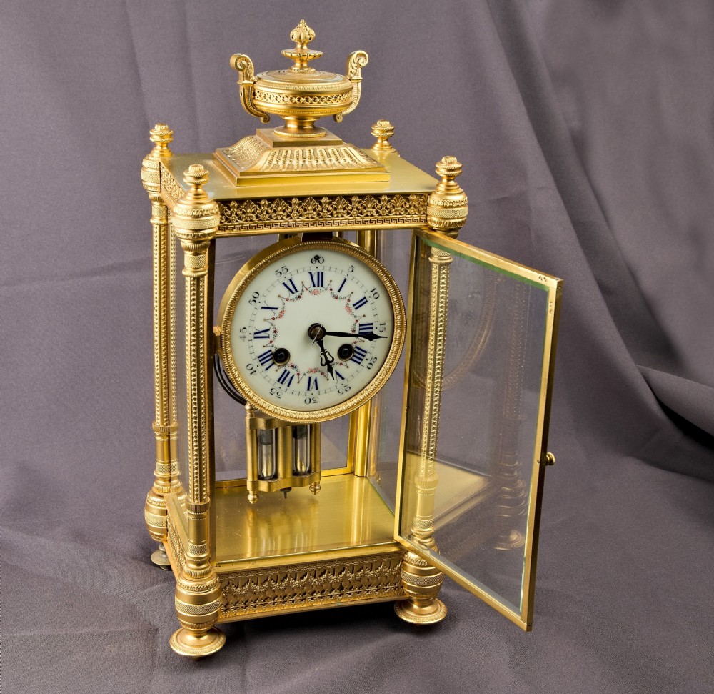 A French Neoclassical gilt brass four-glass mantel clock featuring an urn finial, fluted corner columns, an enamel dial with Roman numerals, and a visible mercury pendulum. View IX