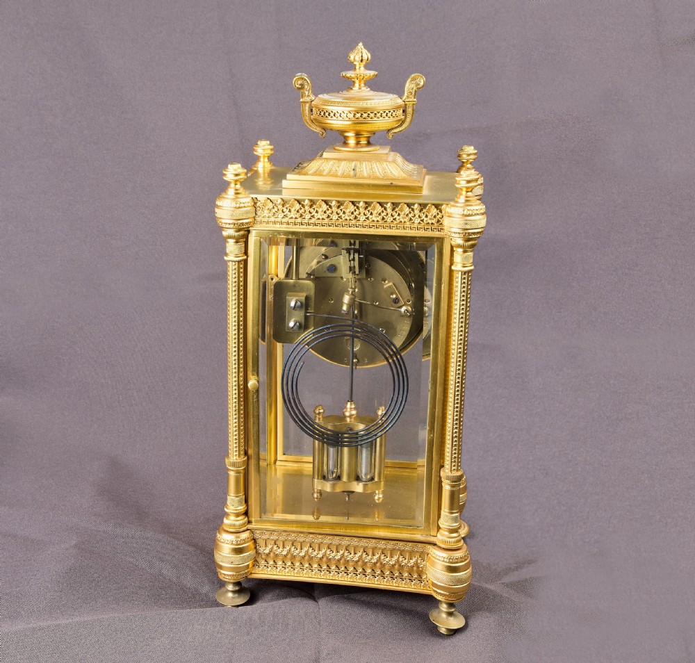A French Neoclassical gilt brass four-glass mantel clock featuring an urn finial, fluted corner columns, an enamel dial with Roman numerals, and a visible mercury pendulum. View X