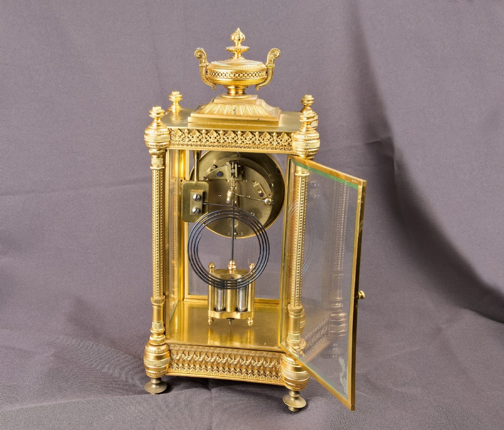 A French Neoclassical gilt brass four-glass mantel clock featuring an urn finial, fluted corner columns, an enamel dial with Roman numerals, and a visible mercury pendulum. View XI
