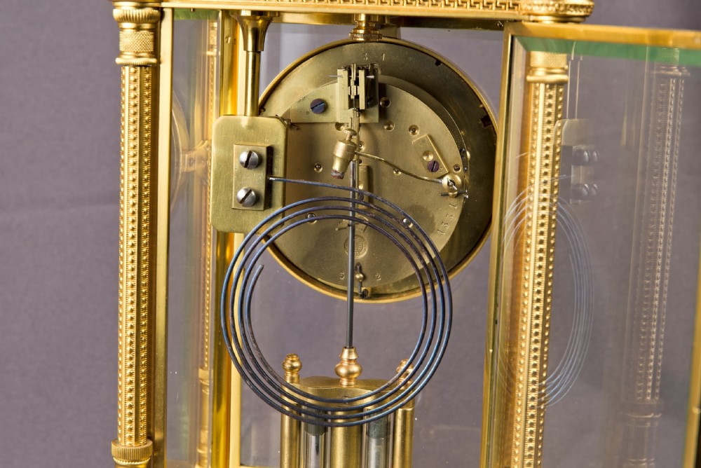 A French Neoclassical gilt brass four-glass mantel clock featuring an urn finial, fluted corner columns, an enamel dial with Roman numerals, and a visible mercury pendulum. View XII