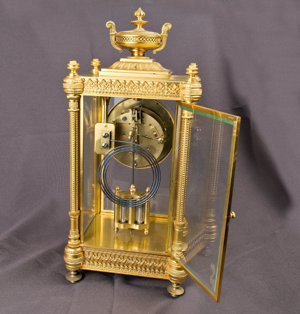 A French Neoclassical gilt brass four-glass mantel clock featuring an urn finial, fluted corner columns, an enamel dial with Roman numerals, and a visible mercury pendulum. View XIII