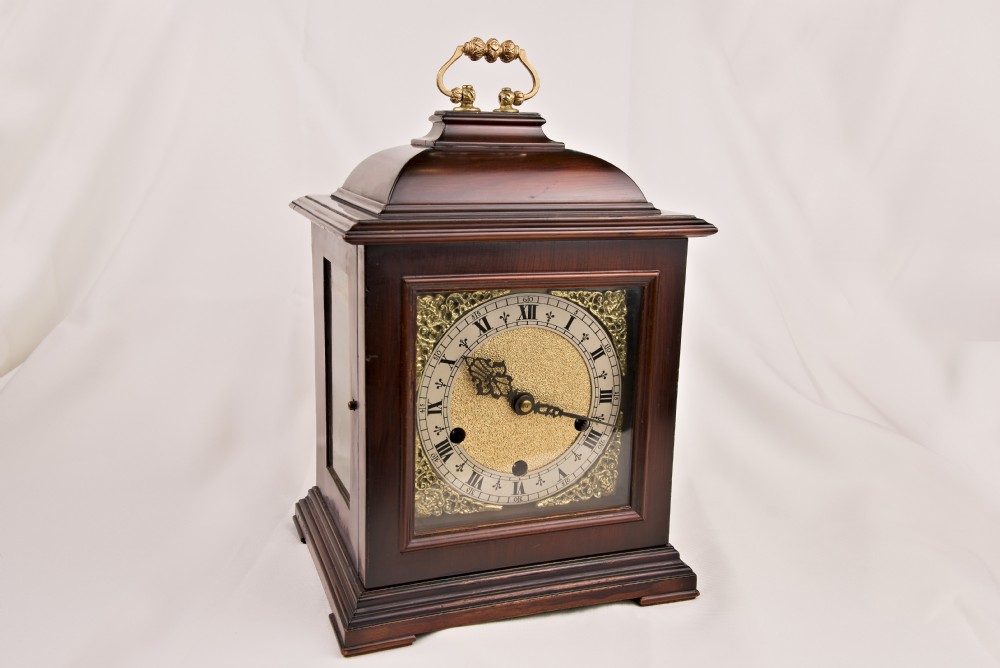 A dark mahogany bracket clock featuring a brass carrying handle on top, side glass panels, and an ornate brass dial with a silvered chapter ring.