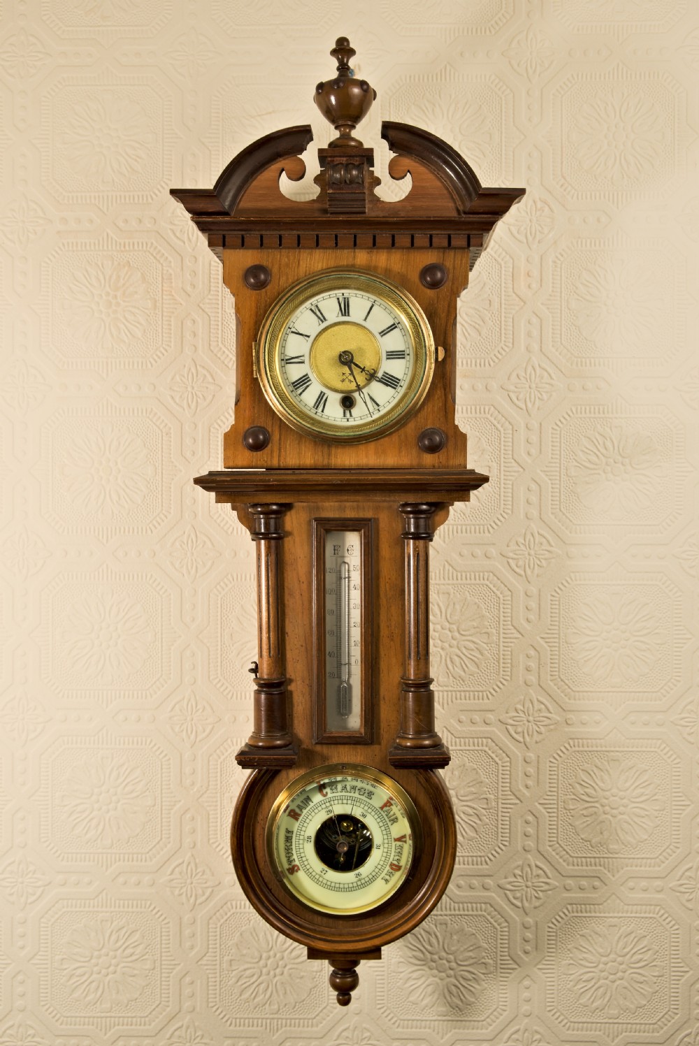 A wall-hung antique walnut clock, thermometer, and aneroid barometer companion set featuring carved pediment ornamentation and brass-rimmed dials.