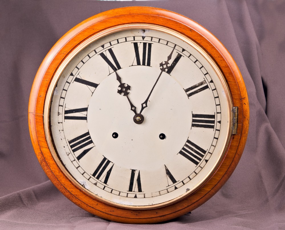 A circular antique wall clock featuring a golden oak turned surround, a white dial with black Roman numerals, brass bezel, and decorative black hands.