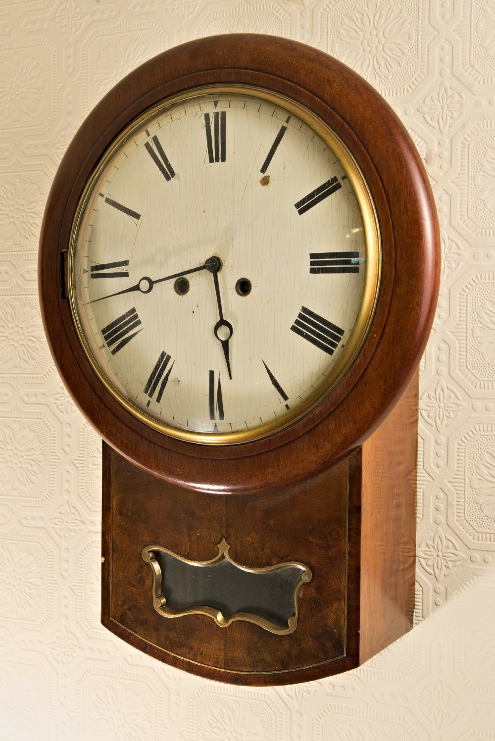 An antique Victorian drop-dial wall clock featuring a circular mahogany frame, cream dial with Roman numerals, and a lower wooden trunk with a brass aperture.