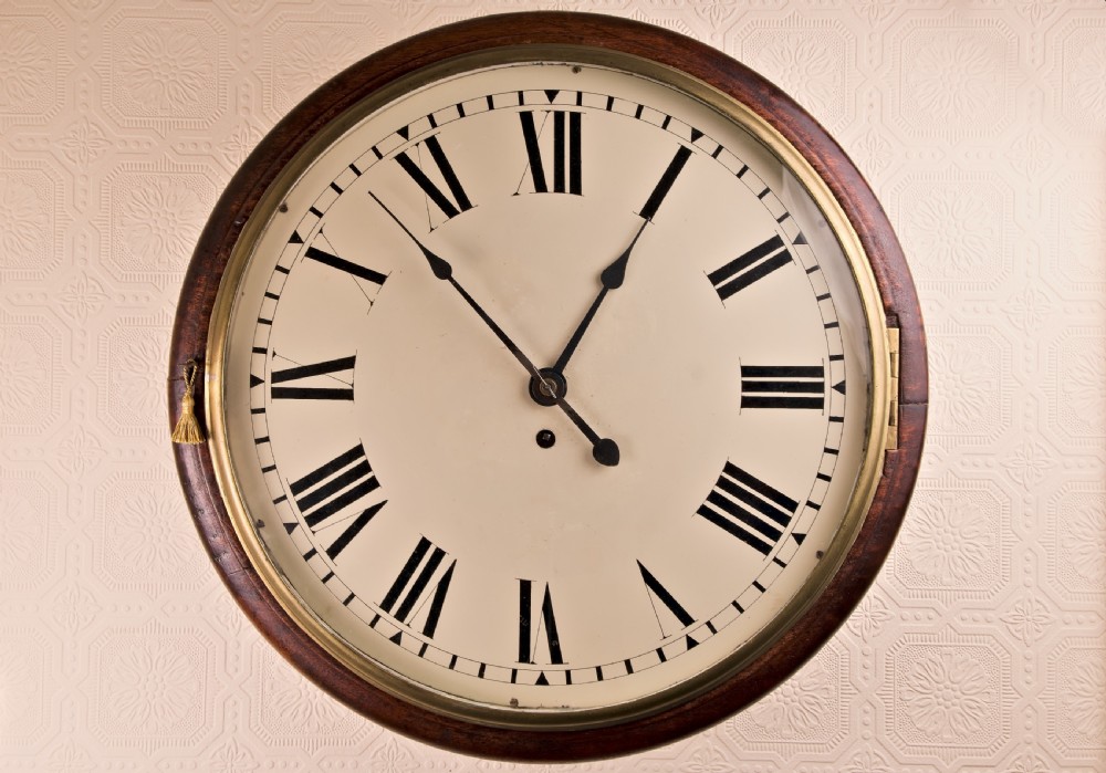 A large circular antique wall clock featuring a dark mahogany surround, turned brass bezel, and a cream dial with black Roman numerals.