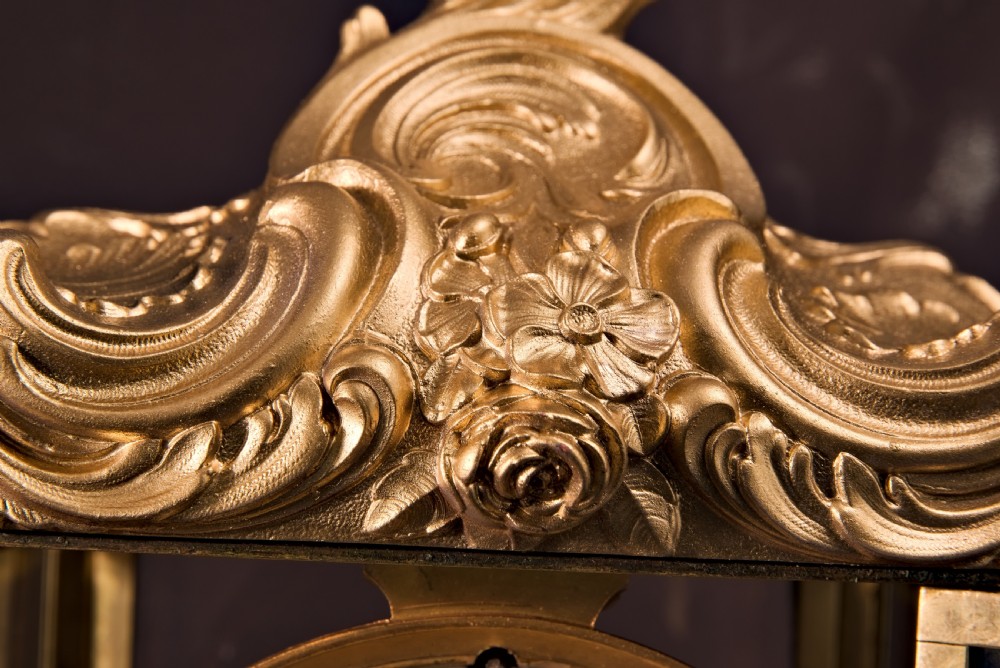 A 19th-century Rococo Revival gilt bronze four-glass mantel clock featuring scrollwork cresting, a white enamel dial, and a visible mercury pendulum. View V