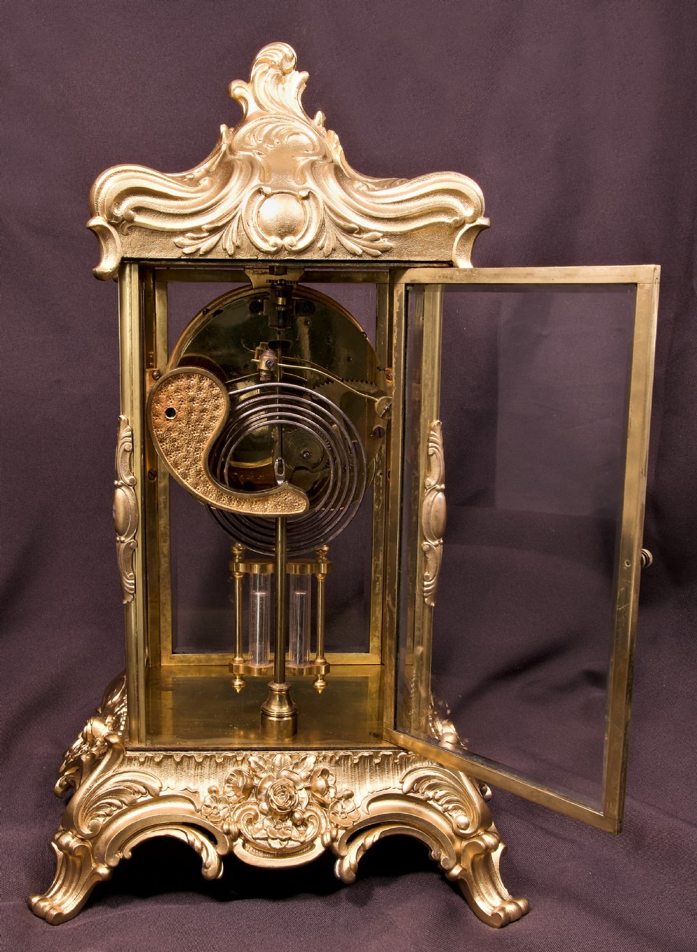 A 19th-century Rococo Revival gilt bronze four-glass mantel clock featuring scrollwork cresting, a white enamel dial, and a visible mercury pendulum. View VII