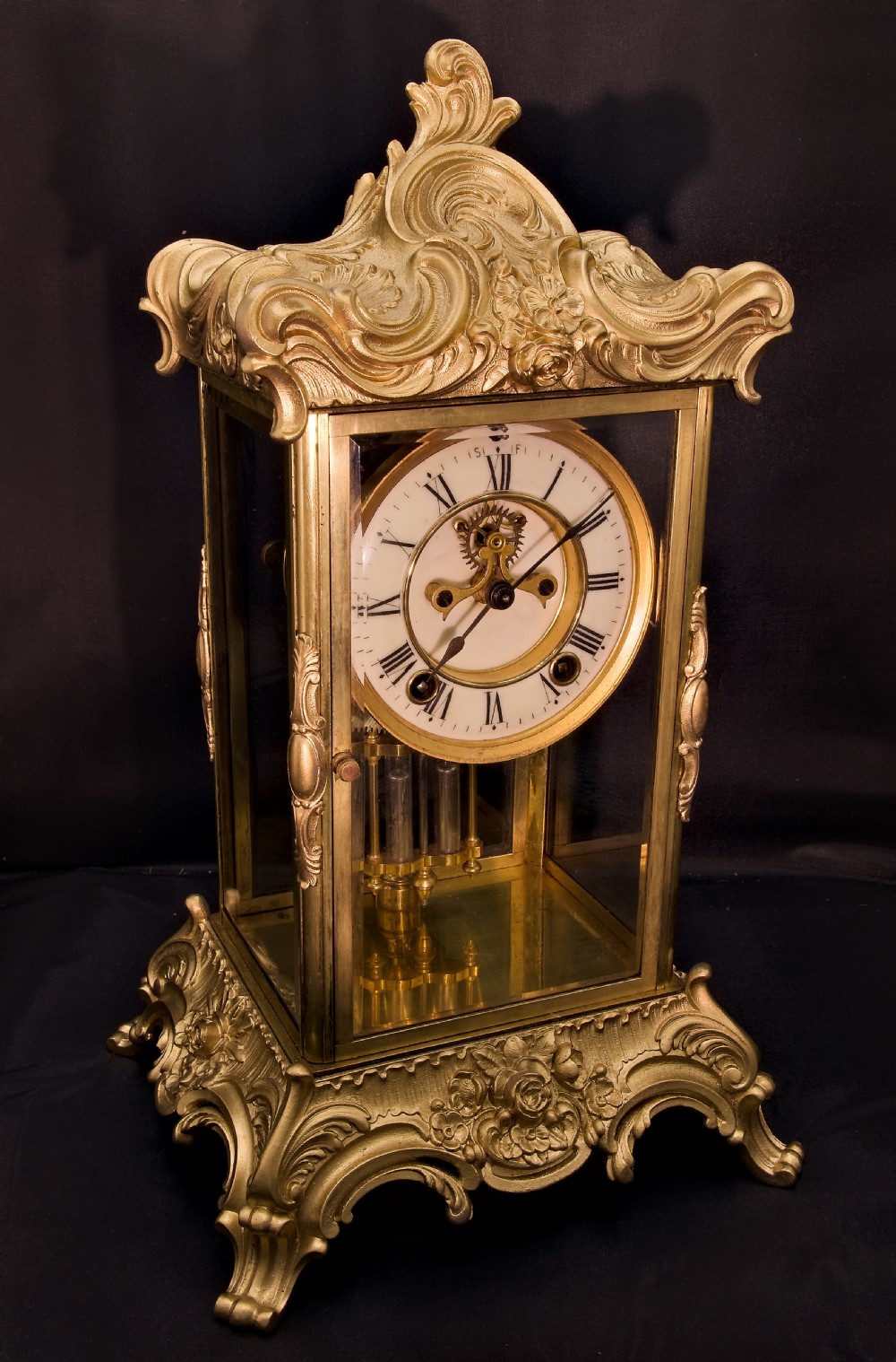 A 19th-century Rococo Revival gilt bronze four-glass mantel clock featuring scrollwork cresting, a white enamel dial, and a visible mercury pendulum. View XI