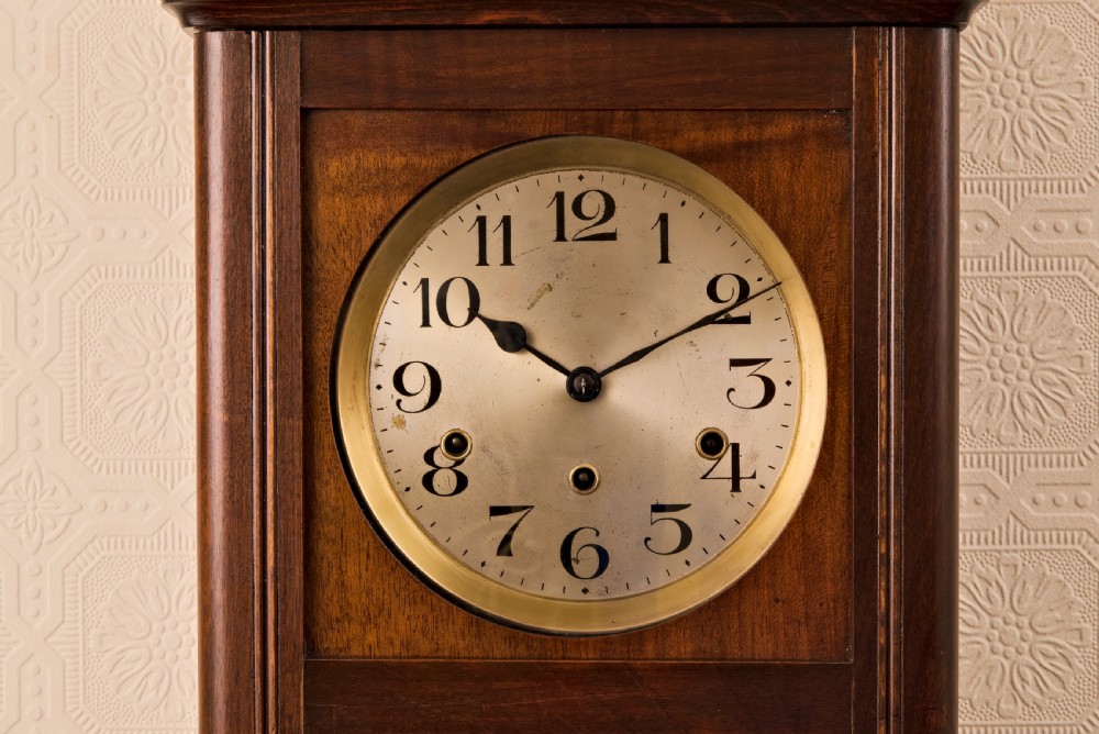 A dark oak Arts and Crafts wall clock featuring a carved shell crest, round dial with Arabic numerals, and a glazed door displaying a brass pendulum. View II