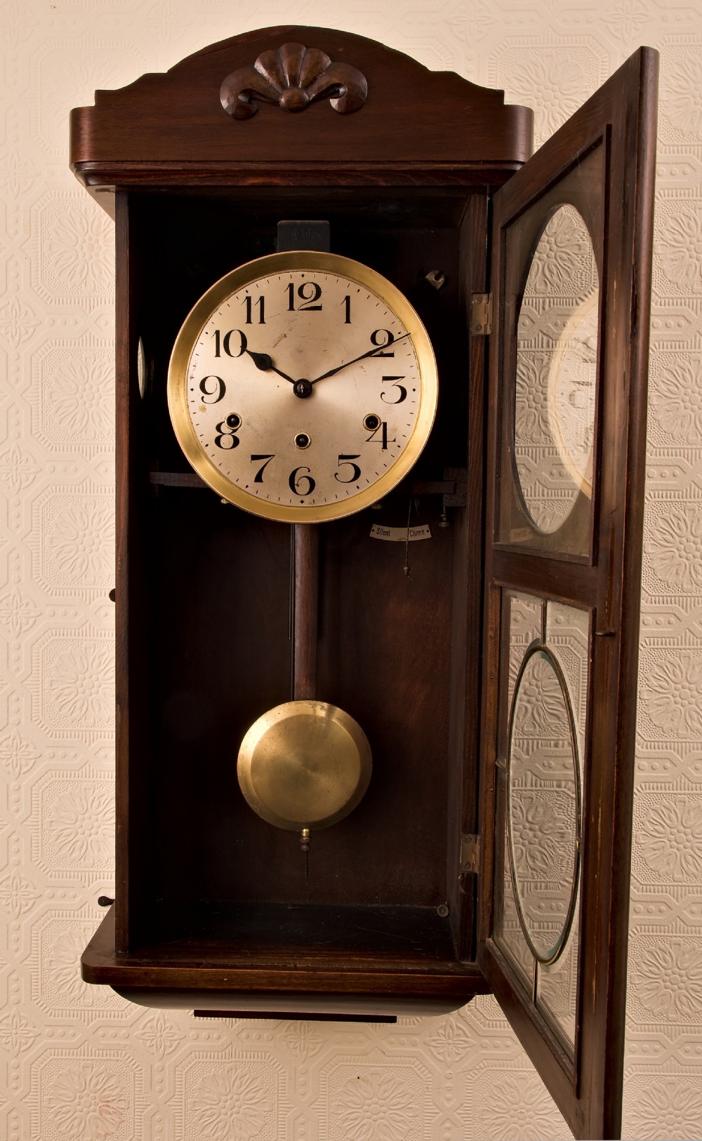 A dark oak Arts and Crafts wall clock featuring a carved shell crest, round dial with Arabic numerals, and a glazed door displaying a brass pendulum. View IV
