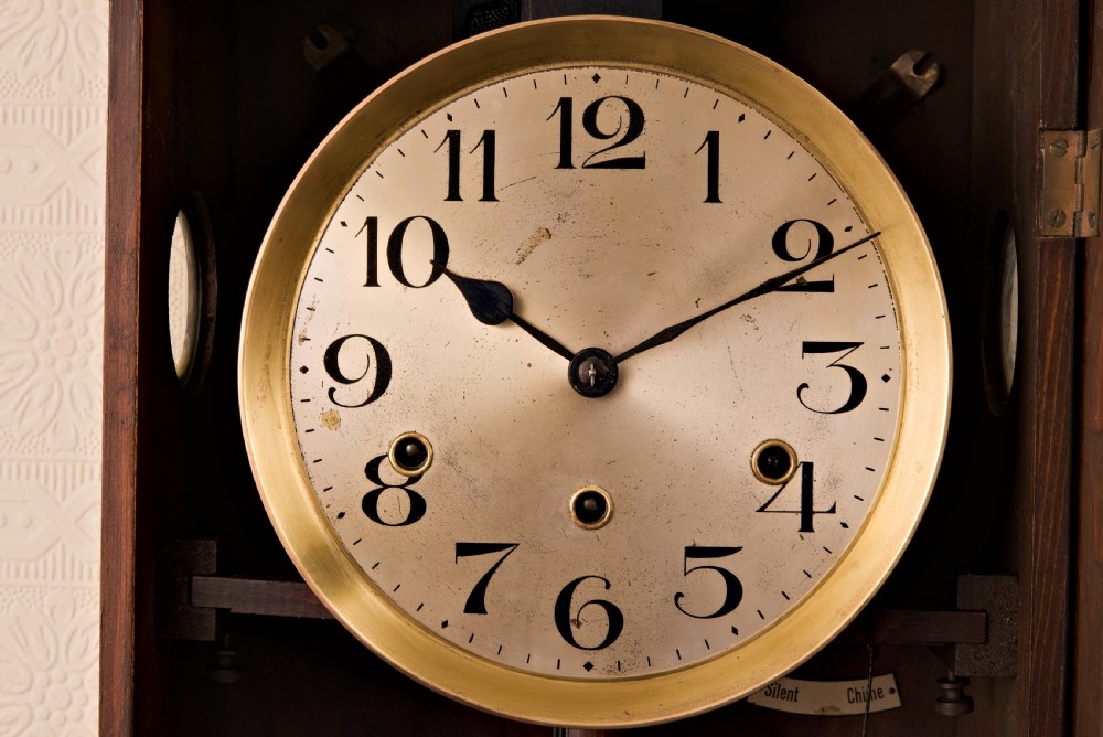 A dark oak Arts and Crafts wall clock featuring a carved shell crest, round dial with Arabic numerals, and a glazed door displaying a brass pendulum. View V