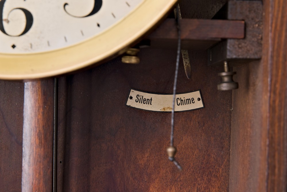 A dark oak Arts and Crafts wall clock featuring a carved shell crest, round dial with Arabic numerals, and a glazed door displaying a brass pendulum. View VI