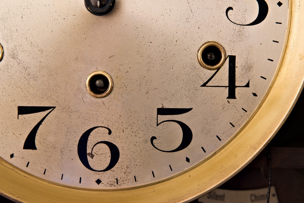 A dark oak Arts and Crafts wall clock featuring a carved shell crest, round dial with Arabic numerals, and a glazed door displaying a brass pendulum. View VII