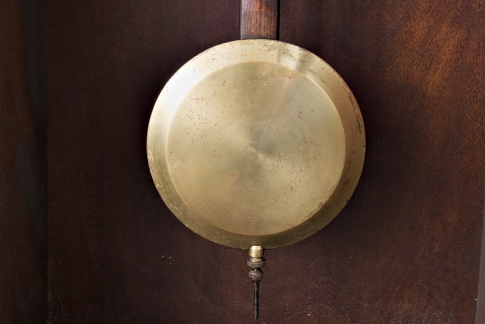 A dark oak Arts and Crafts wall clock featuring a carved shell crest, round dial with Arabic numerals, and a glazed door displaying a brass pendulum. View VIII