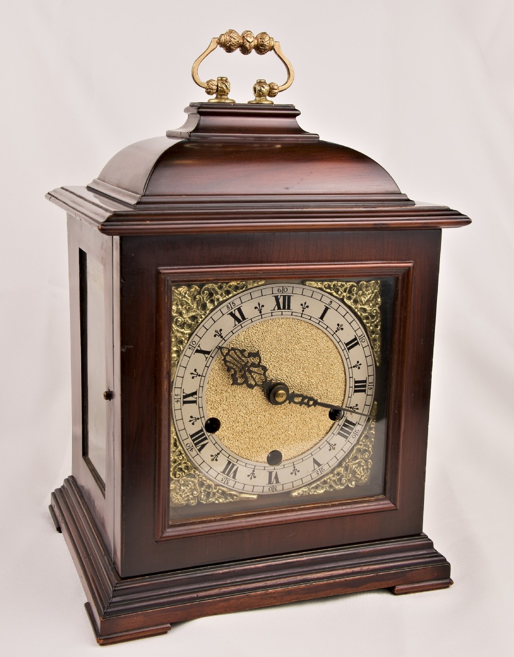 A dark mahogany wooden bracket mantel clock featuring a brass dial with Roman numerals, cast brass spandrels, glass side panels, and a brass carrying handle on top.