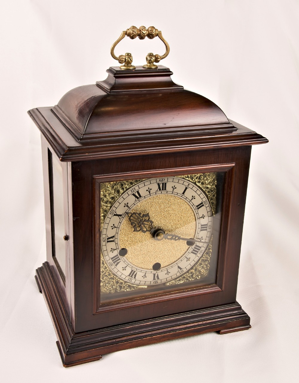 A dark mahogany wooden bracket mantel clock featuring a brass dial with Roman numerals, cast brass spandrels, glass side panels, and a brass carrying handle on top. View X